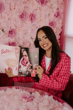 Smiling woman in pink houndstooth blazer holds magazine and softball poster against pink flower wall.
