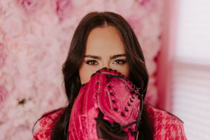 Woman with dark hair holds a pink baseball glove over her face against a floral pink background.