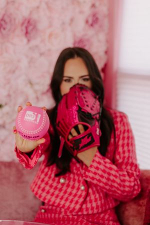 Woman in pink houndstooth outfit holding a pink baseball glove and ball against a floral background.