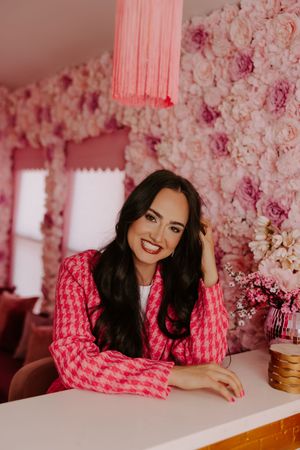 Woman in pink houndstooth blazer smiling at camera against a floral pink rose wall backdrop.