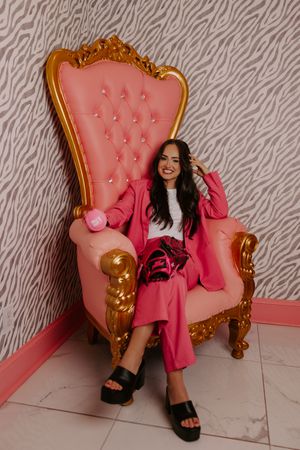 Smiling woman in pink pantsuit sitting on ornate gold and pink throne chair against zebra print wall.
