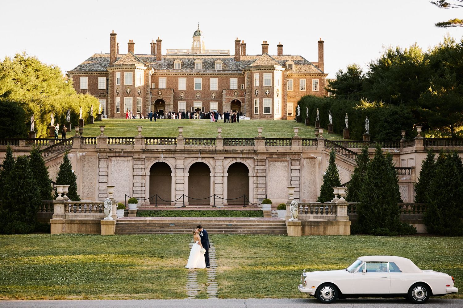 Interracial couple in wedding attire embracing in a lush garden, surrounded by white flowers at sunset.