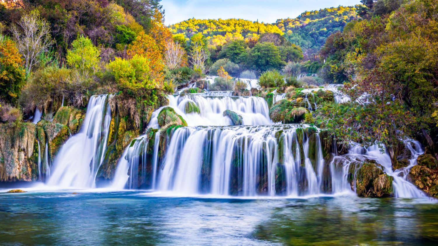 Stunning Krka National Park waterfalls cascade over lush green rocks surrounded by colorful autumn forest in Croatia.