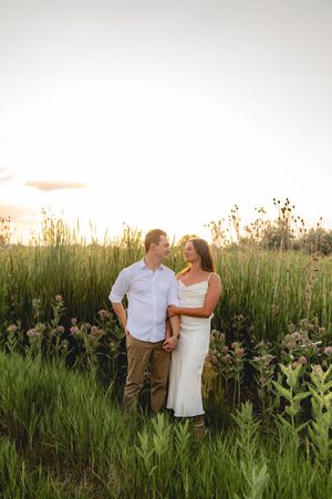 Couple holding hands in a wildflower field at golden hour sunset, man in blue shirt and woman in white dress.