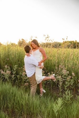 Couple embracing in a lush green field at golden hour, woman in white dress lifted by man in white shirt.