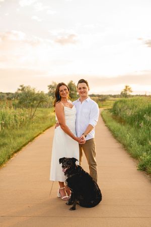 Couple holding hands with black dog on scenic path at golden hour sunset, surrounded by lush green grass.
