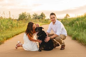 Couple crouching on a scenic path with their black dog at golden hour, surrounded by lush green grass.