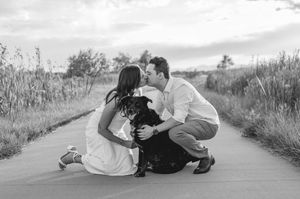 Black and white photo of a couple kissing while crouching with their black dog on a scenic pathway.