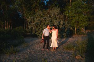 Couple in wedding attire walking hand in hand on a sandy path surrounded by lush green trees at dusk.