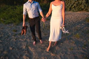 Barefoot couple holding hands on sandy beach, woman in white dress carrying shoes, man in blue shirt holding sandals.