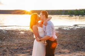 Couple sharing a romantic kiss on a sandy beach at golden sunset with a shimmering lake in the background.