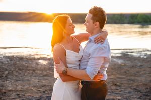 Couple embracing at golden hour sunset by a river, woman in white dress and man in light shirt gazing at each other.