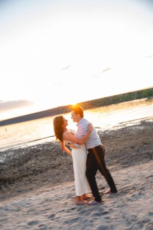 Couple embracing on sandy beach at golden sunset, romantic engagement photo with warm sunlight glowing behind them.