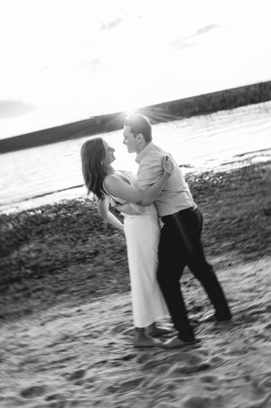 Black and white photo of a couple dancing romantically on a sandy beach at sunset near the water.