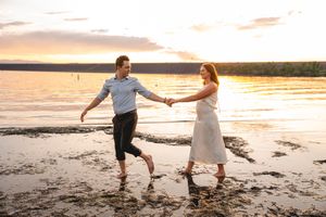 Couple holding hands walking barefoot on a sandy beach shore at golden sunset by a calm lake.