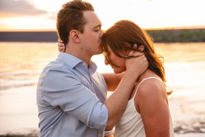 Romantic couple sharing a tender kiss at sunset by the water, man in blue shirt embracing woman with red hair.