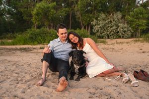 Couple sitting on sandy beach with black dog, surrounded by lush green trees at sunset.