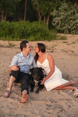 Couple sitting on sandy beach with black dog, smiling at each other, trees in background, golden hour light.