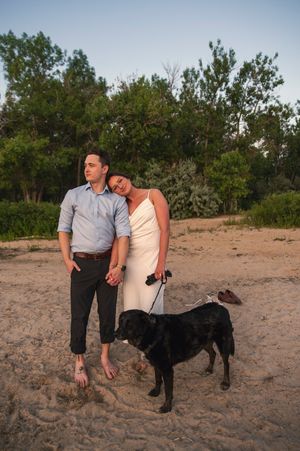Couple in white dress and blue shirt standing barefoot on sandy beach with black Labrador dog at golden hour.