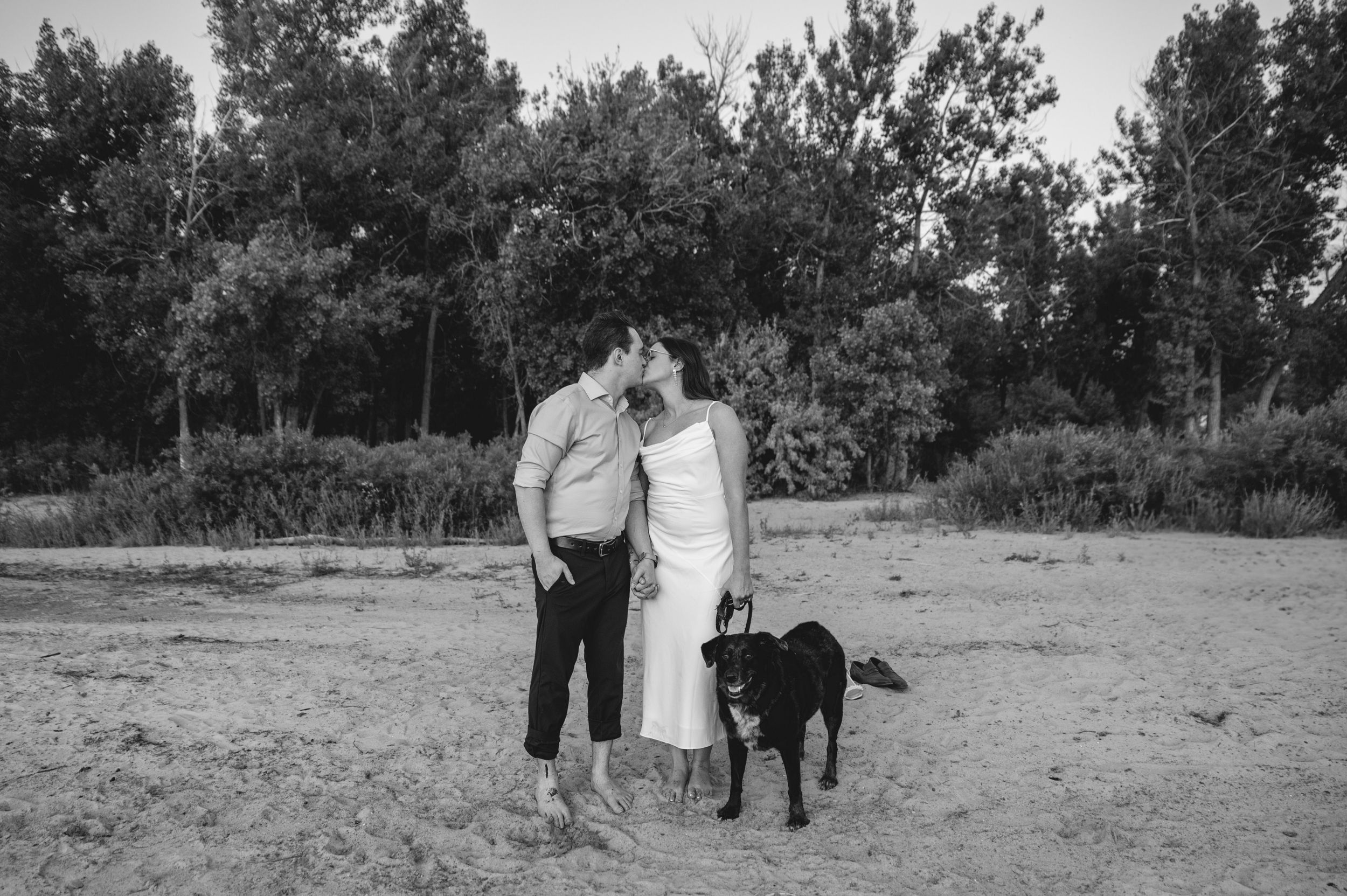 Black and white photo of a couple kissing outdoors with a black Labrador dog standing beside them on sandy ground.