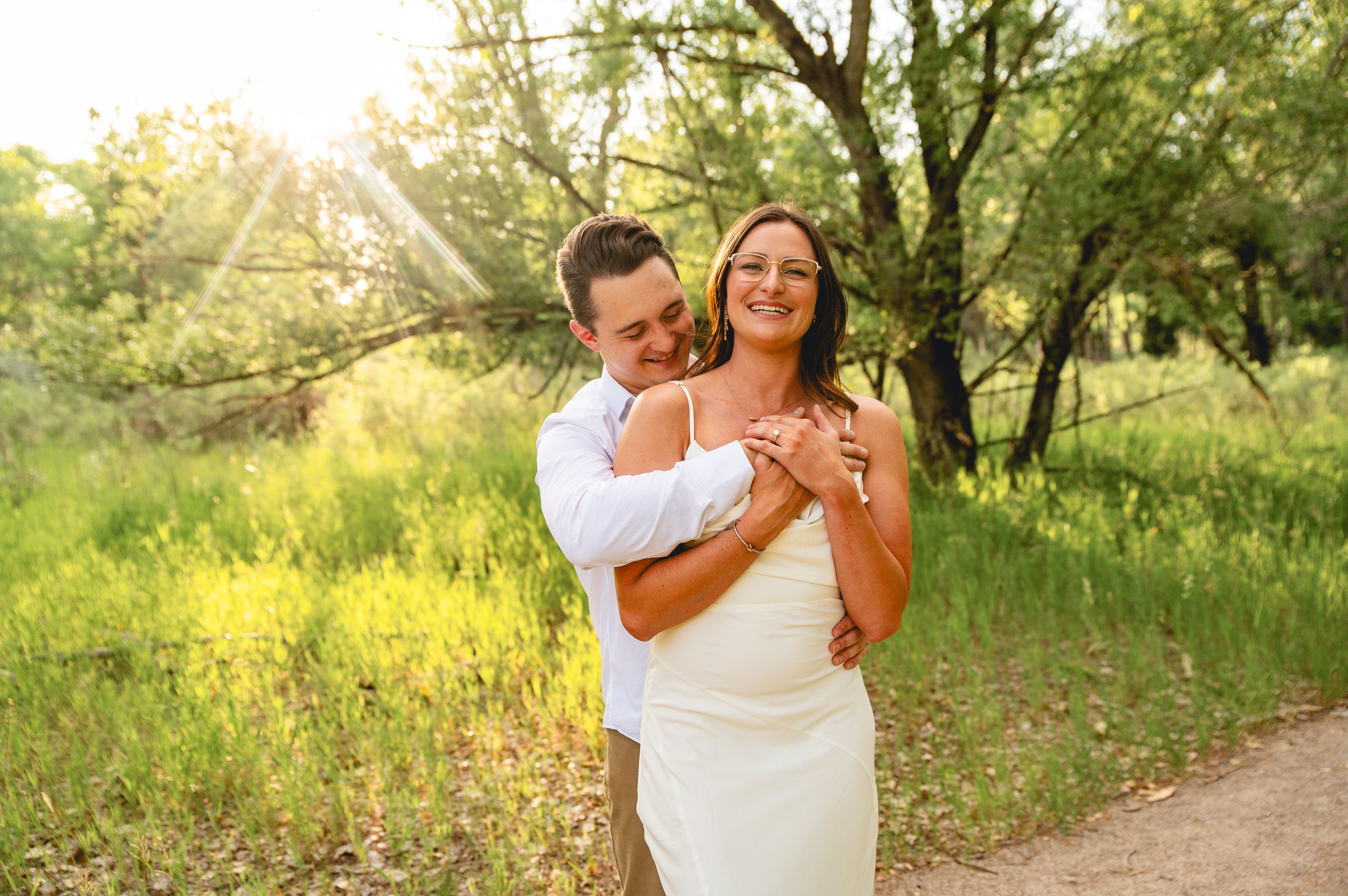 Couple embracing in sunlit forest clearing, bride in white dress smiling as groom hugs her from behind.