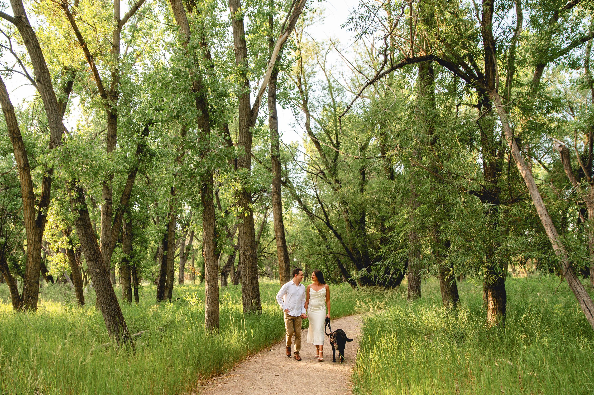 Couple walking dog on scenic forest path surrounded by tall green trees and lush grass.