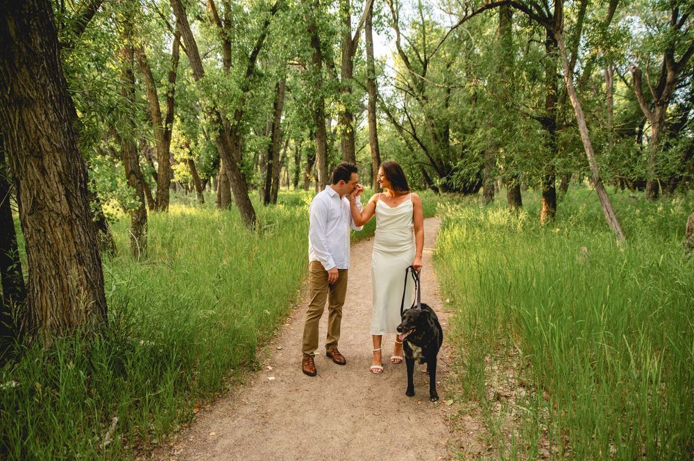 Couple walking black dog on wooded trail, woman in white dress and man in casual attire enjoying nature.
