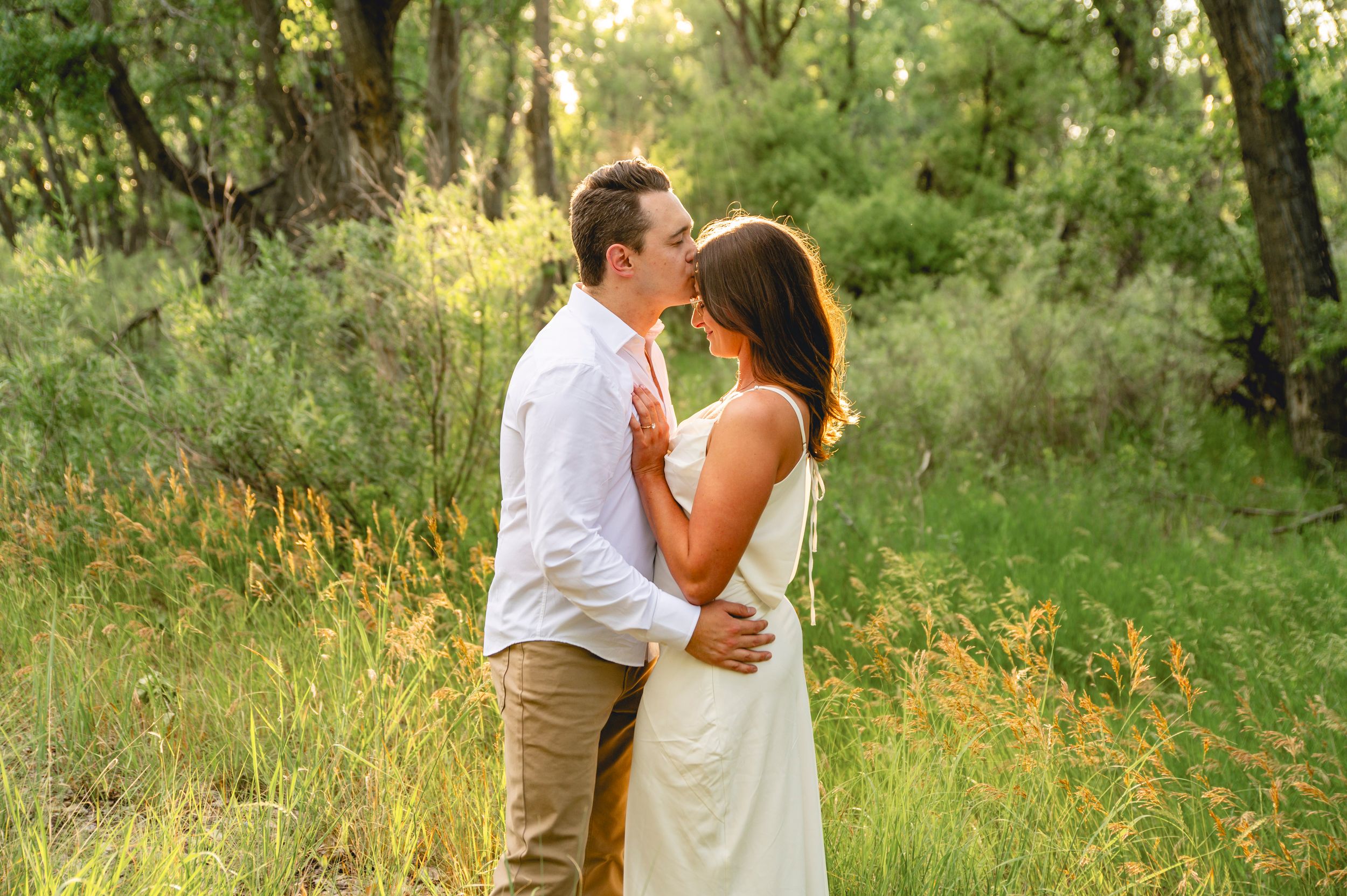 Couple embracing and kissing in a sunlit forest meadow, man in white shirt and woman in white dress.
