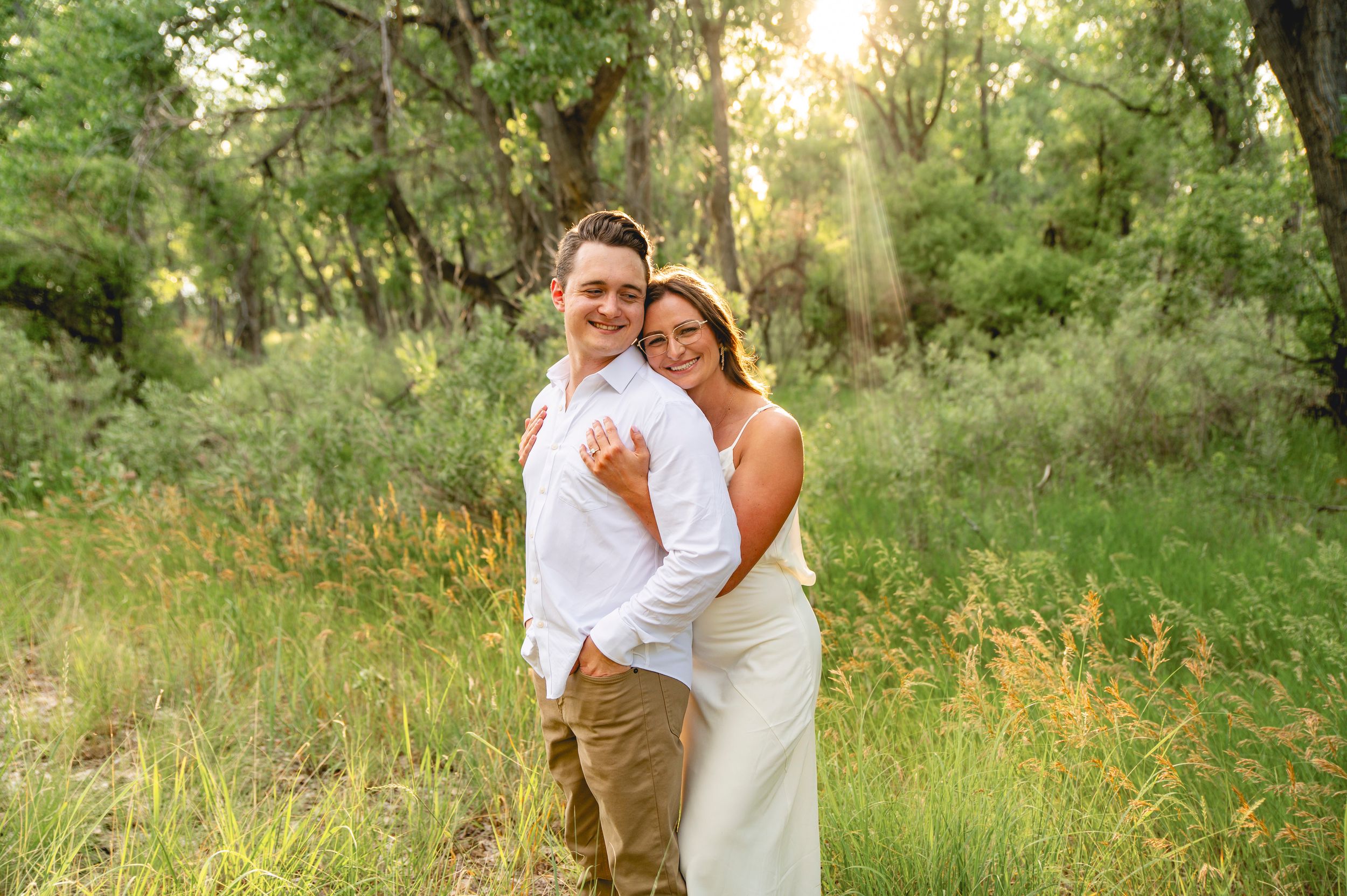 A couple poses in a lush green forest, the woman in white embracing the man in a white shirt and khaki pants.