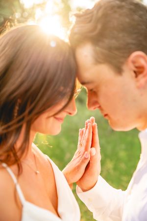 Couple touching foreheads and pressing palms together in golden sunset light, wearing white wedding attire outdoors.