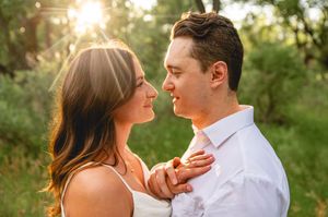 Couple sharing a romantic moment outdoors at golden hour, woman in white dress holding man's tie in lush green setting.