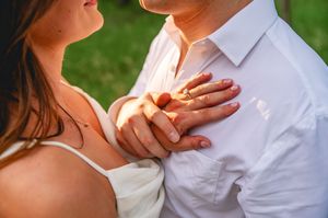 Close-up of a couple holding hands, woman wearing an engagement ring, dressed in white outdoors.