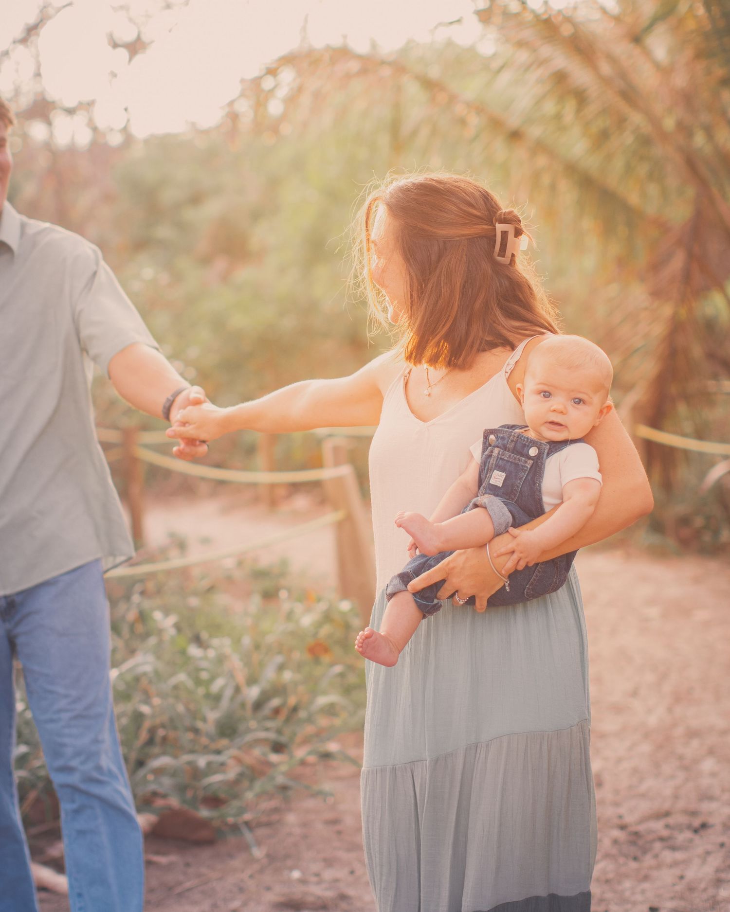 A mother holds her baby while reaching for her husbands hand on a sunlit sandy beach path.