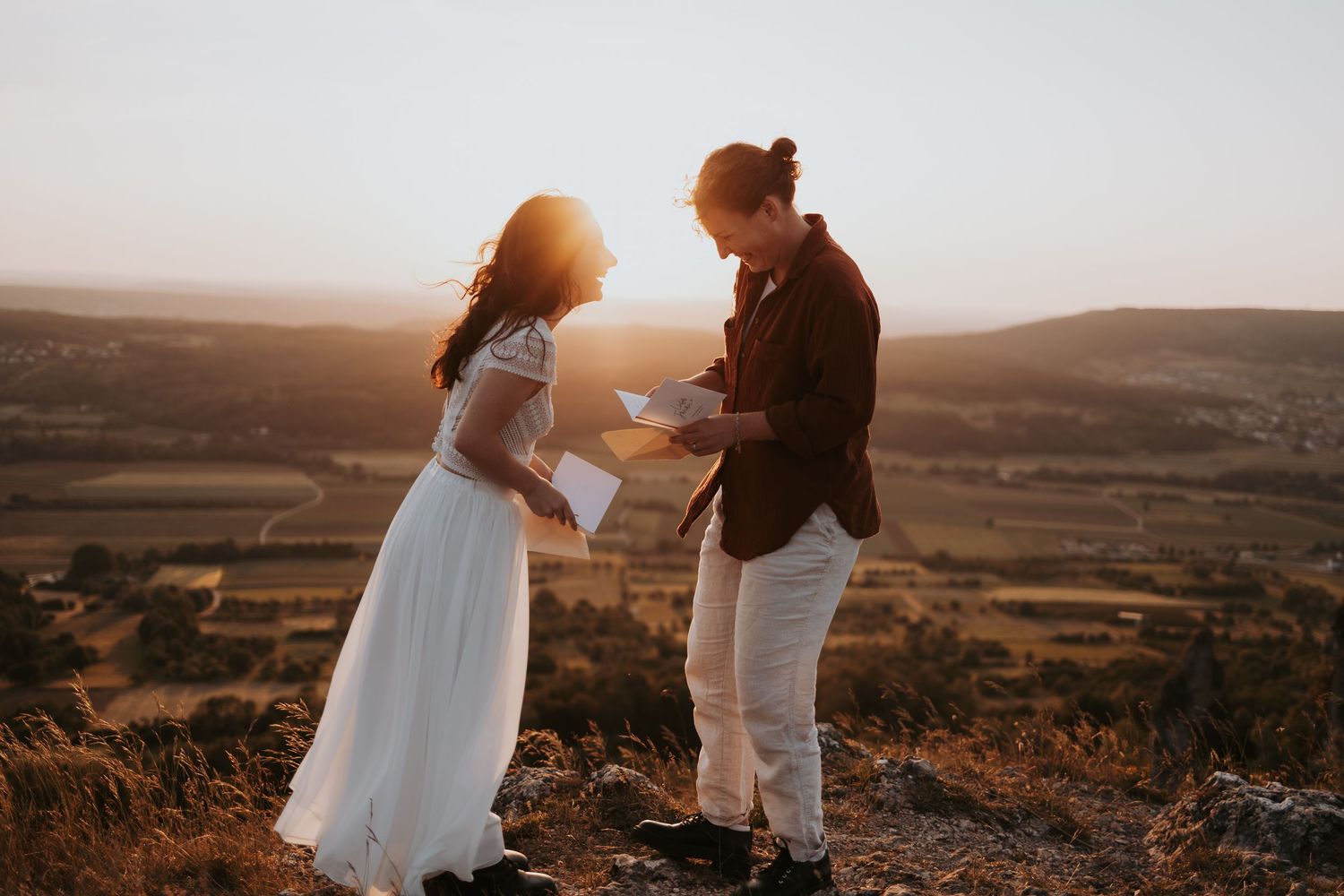 Bride in white tulle gown with long blonde hair walks with groom in tan suit through lush green forest.