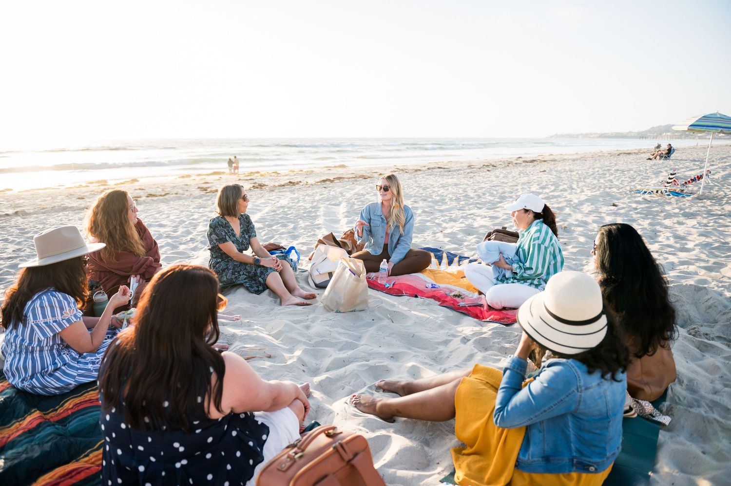 Group of women enjoying a beach picnic gathering on sandy shore at sunset, sitting on colorful blankets together.