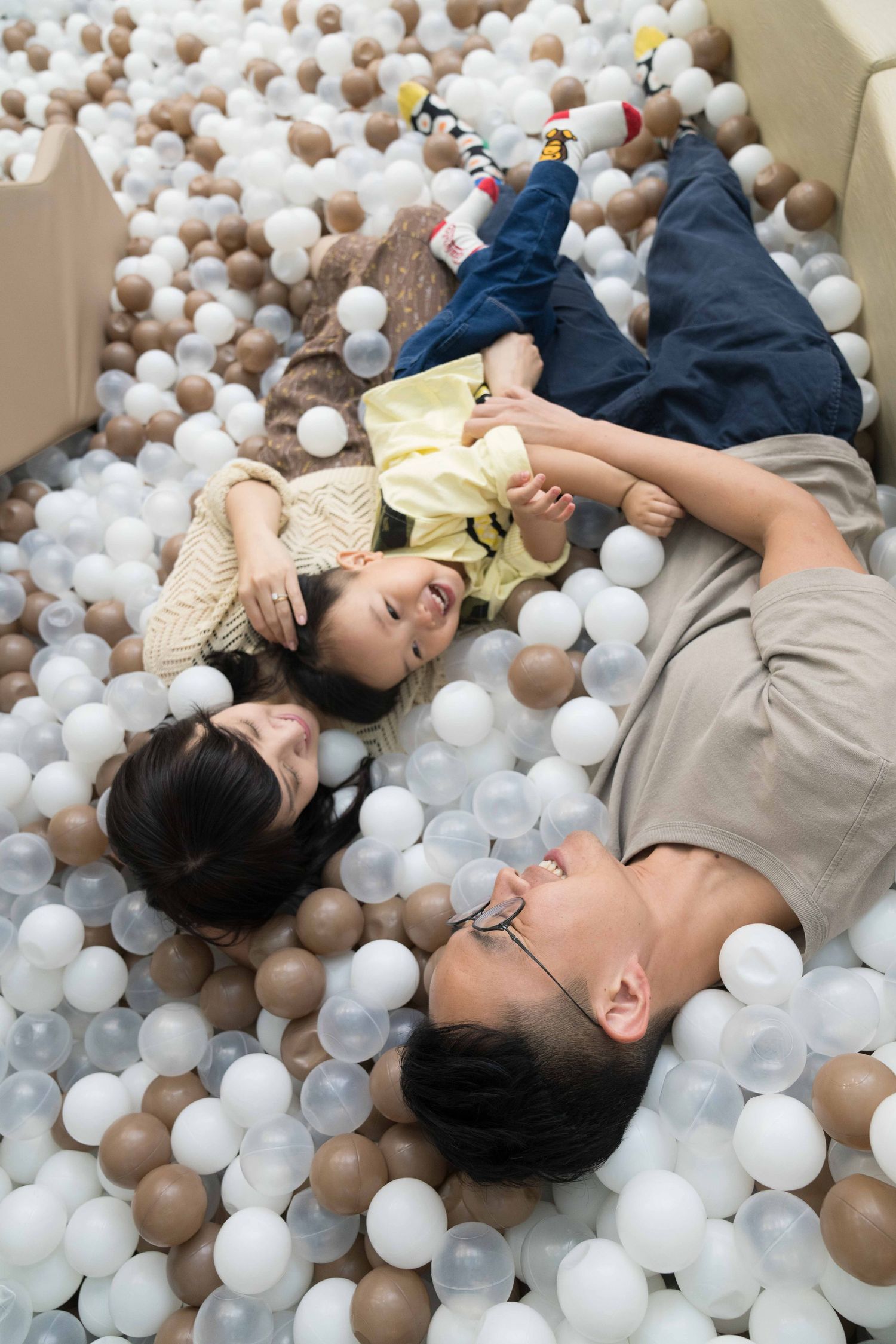 Family of three joyfully playing in a colorful ball pit, lying together and laughing among white and brown plastic balls.
