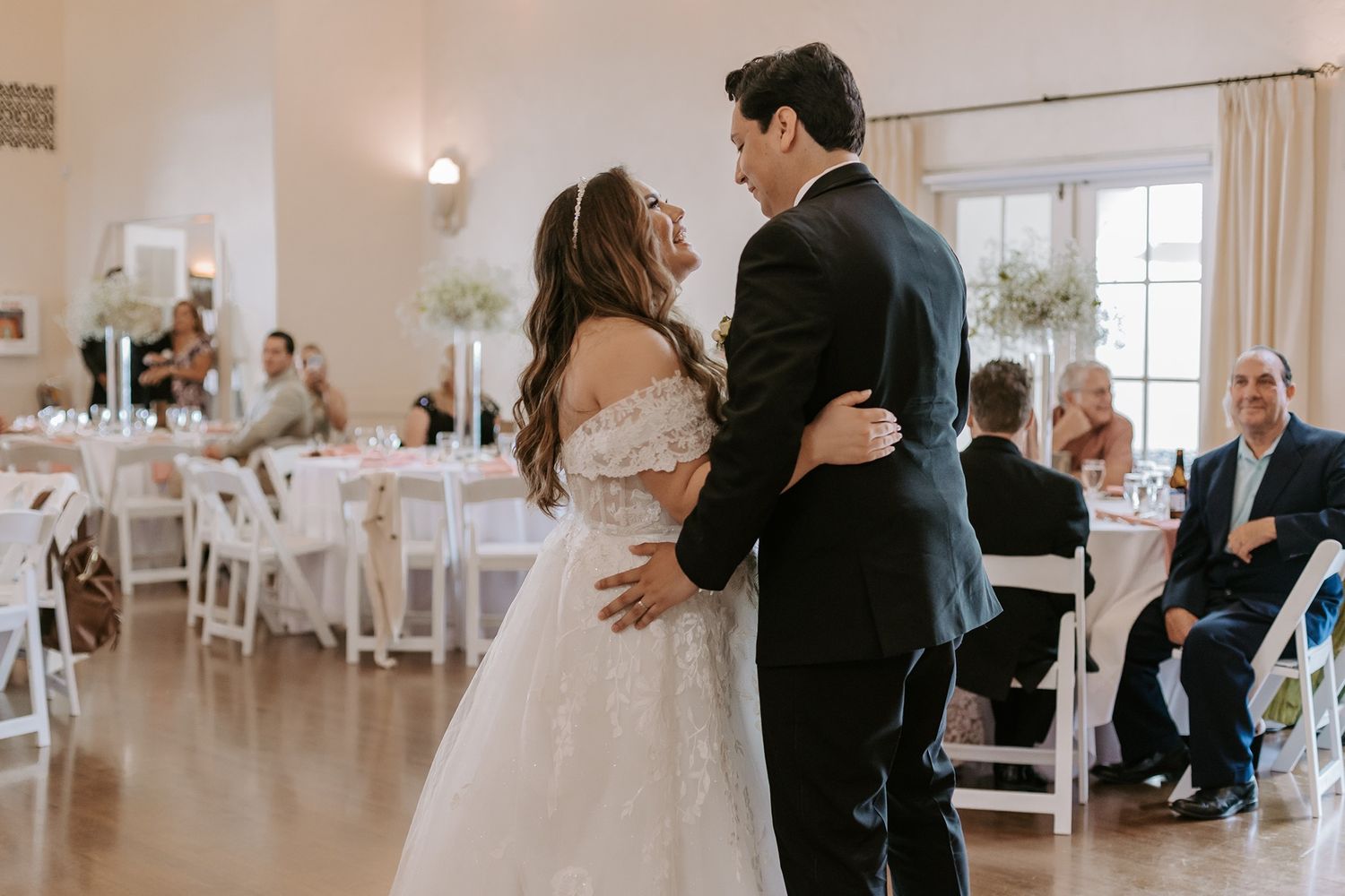 Bride and groom share their first dance at elegant wedding reception with white floral centerpieces and seated guests.