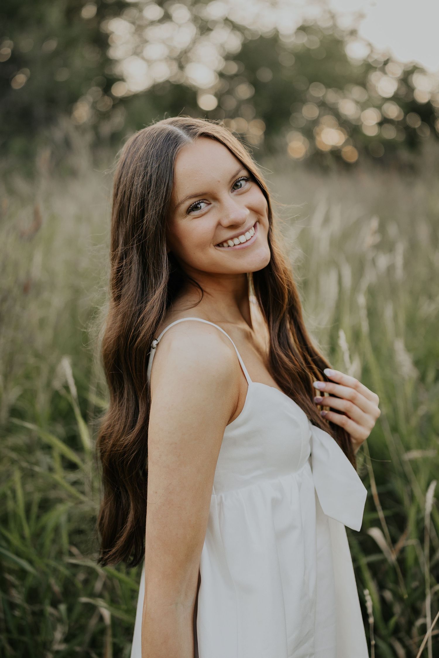 Young woman with long brown hair smiling in a white sundress, standing in a lush green field at golden hour.