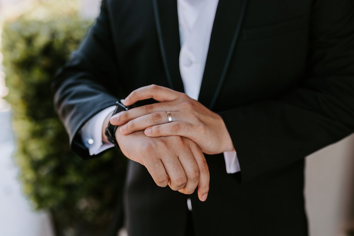Groom in black tuxedo adjusting his cufflinks, wearing a wedding band on his ring finger.