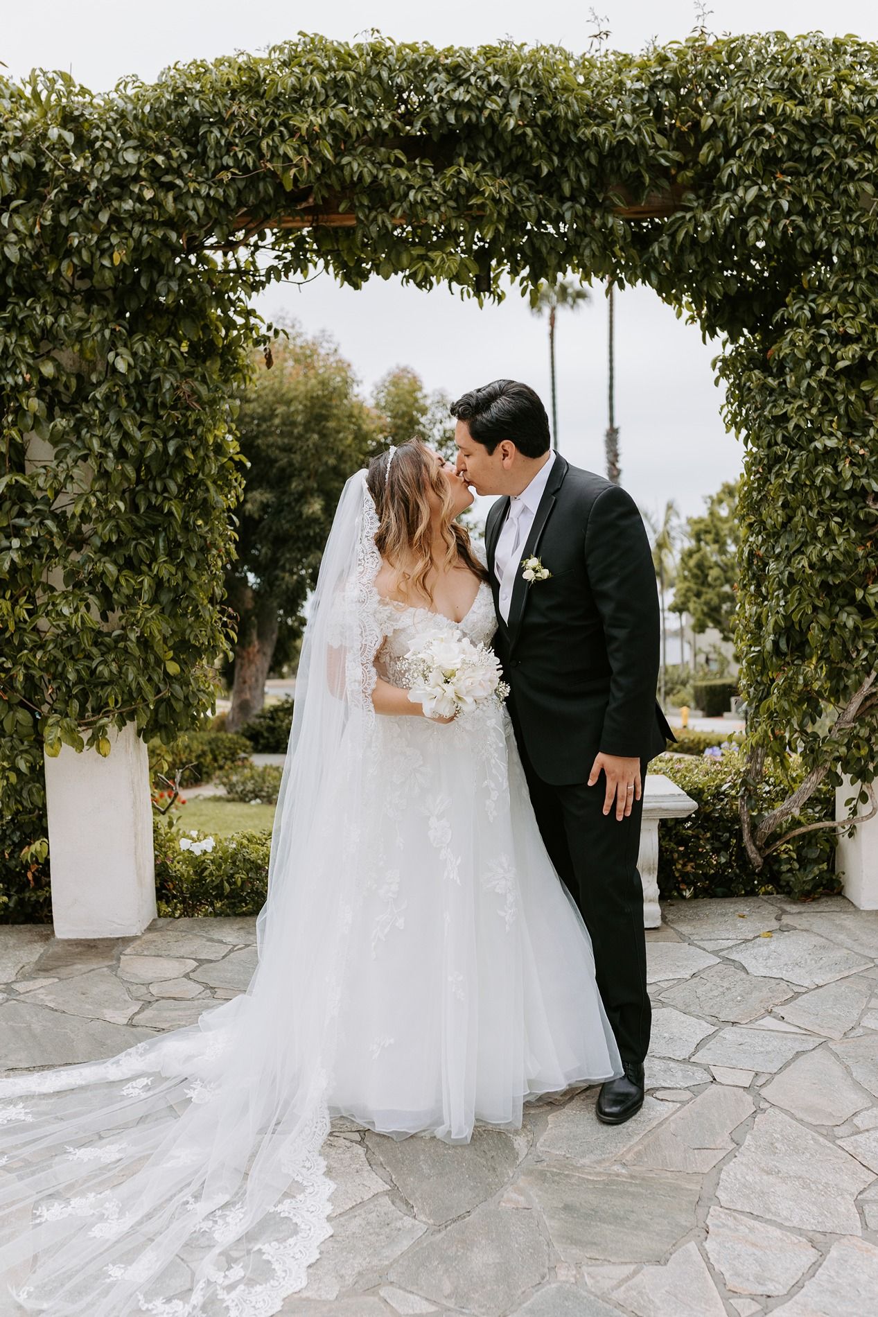 Bride and groom share a kiss under a lush green arch on a stone patio, bride in lace gown with veil and bouquet at The Thursday Club.