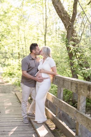 Couple de futurs parents s'embrassant sur un pont de bois dans un sentier boisé lors de leur séance photo de maternité au bord du fleuve