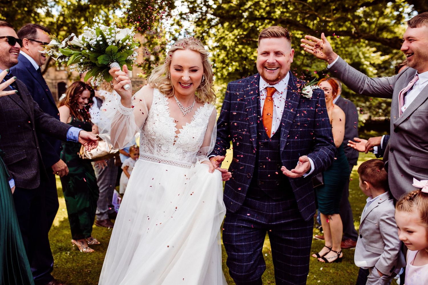 Newlywed couple walking through confetti outside a stone church, surrounded by smiling wedding guests on a sunny day.