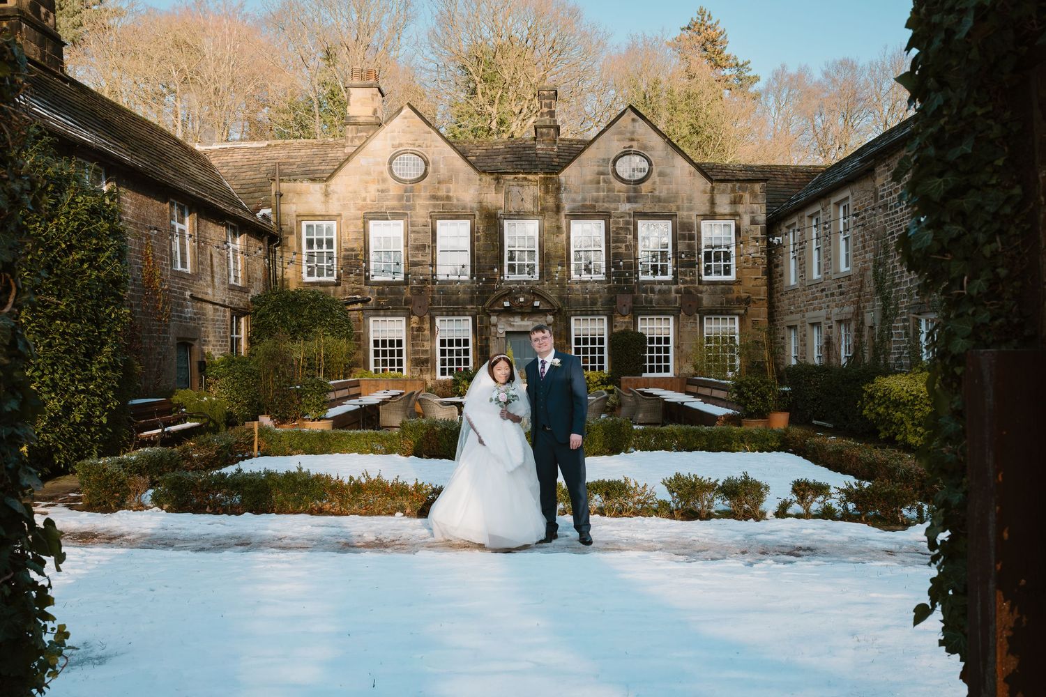 Bride and groom pose in snowy winter garden outside elegant stone manor house wedding venue.