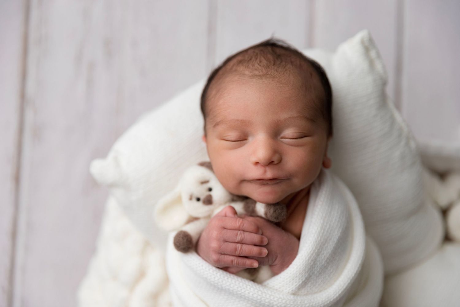 Newborn baby swaddled in white wrap sleeping peacefully while holding a small teddy bear stuffed animal.