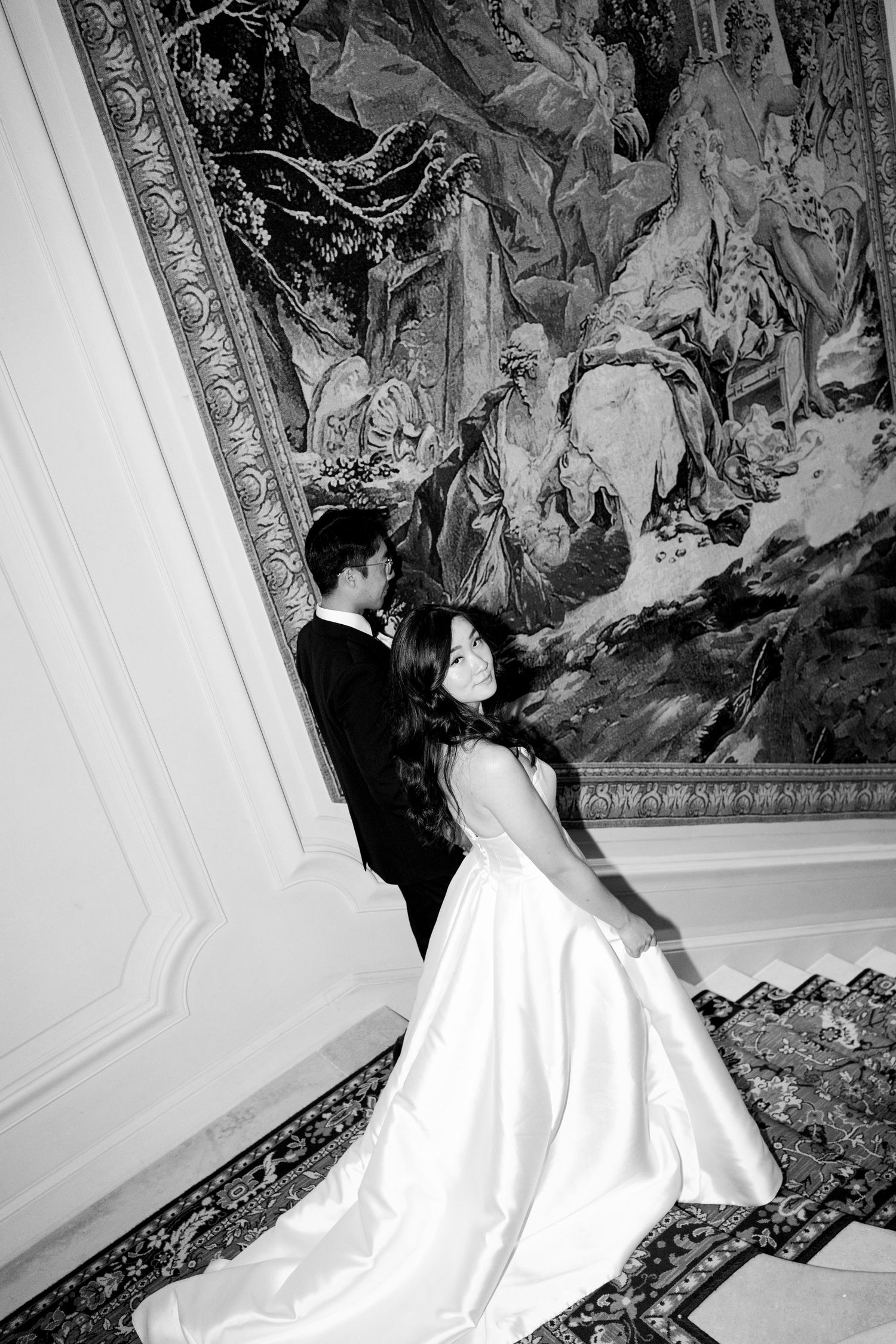 Black and white wedding photo of bride and groom embracing before an ornate tapestry in an elegant venue.