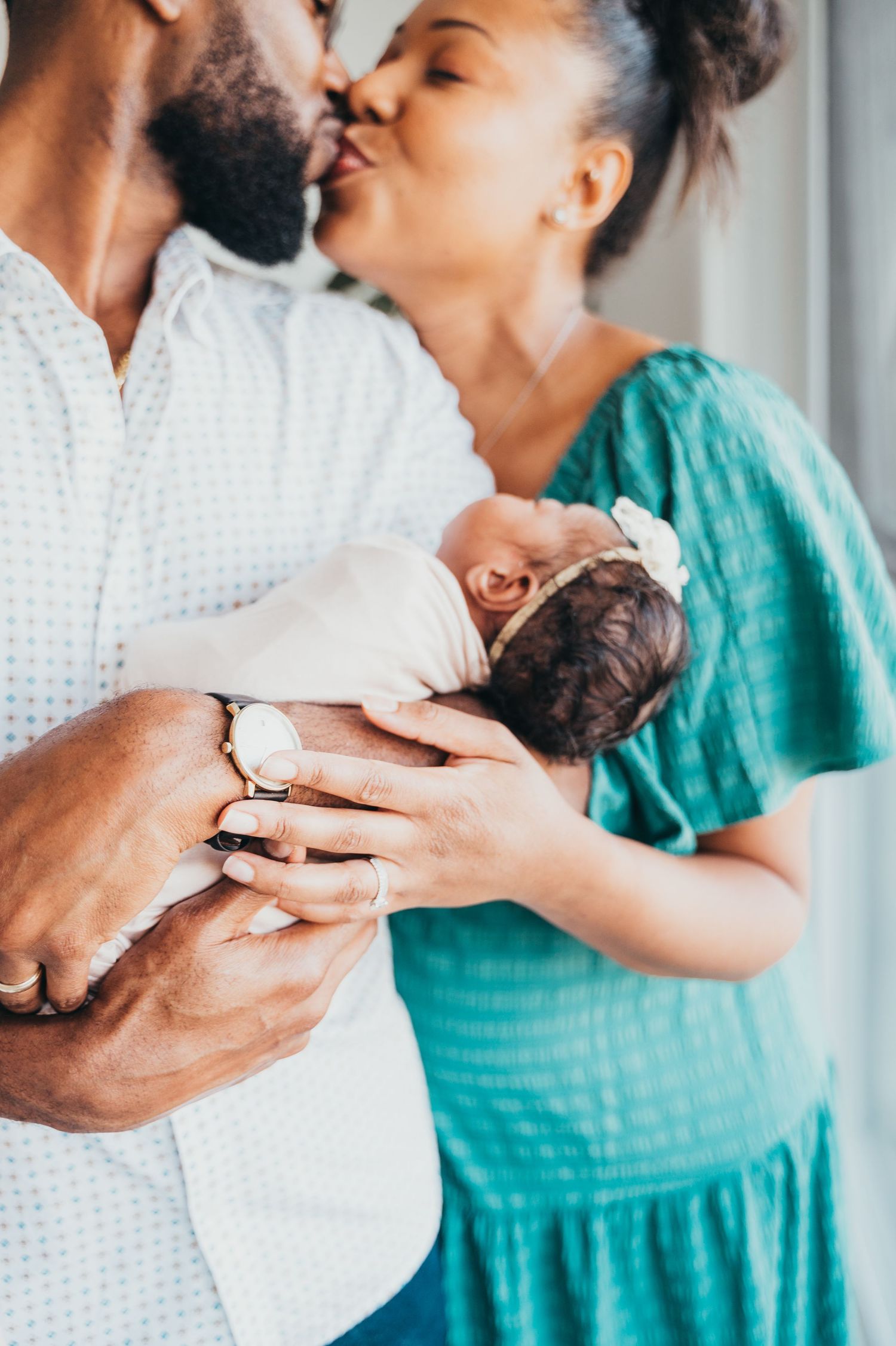 Mom and dad share a kiss while father gently holds their sleeping newborn baby, capturing a tender family moment.