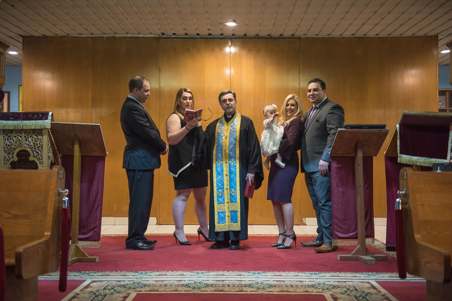 Greek Orthodox priest in blue vestments posing with family after baptism ceremony.