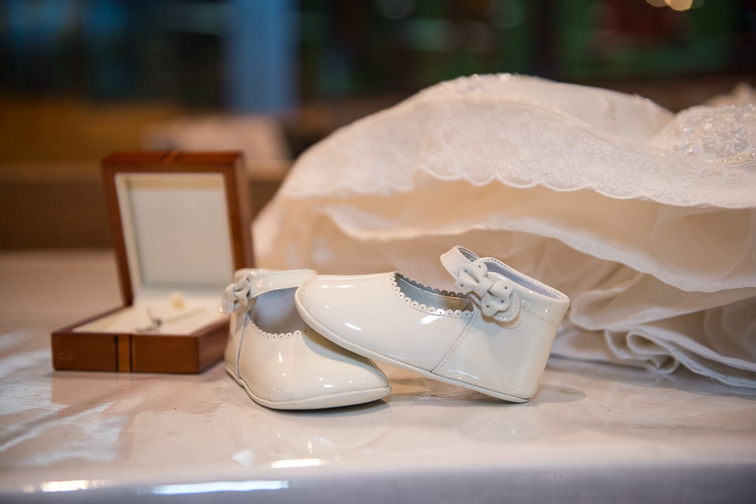 White patent leather baby shoes beside christening gown and jewelry box on white table.