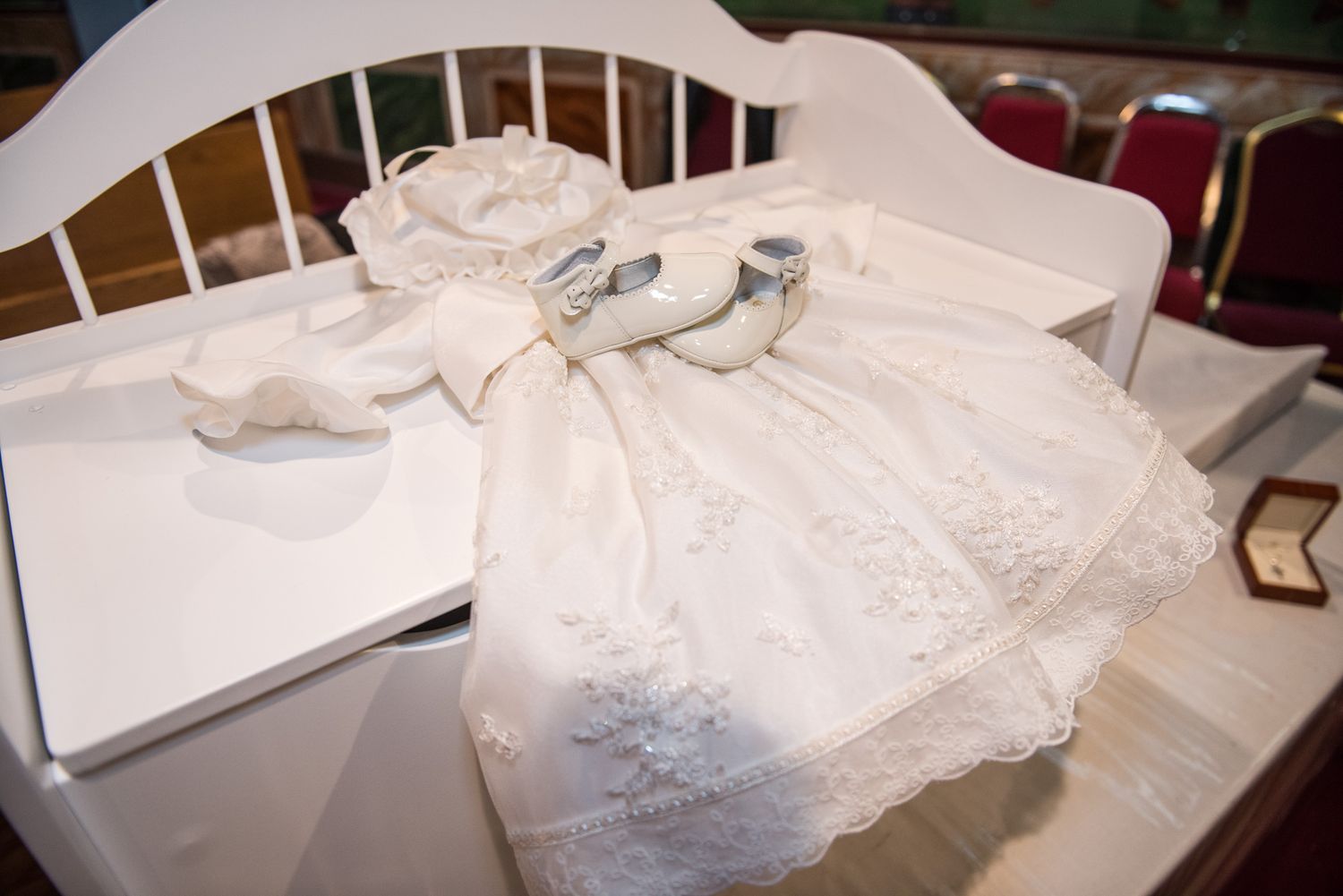 White lace christening gown with matching bonnet and silver shoes displayed on white bench.