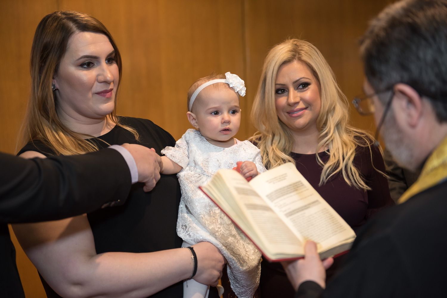Two women holding baby in white christening dress while priest presents holy scripture book.